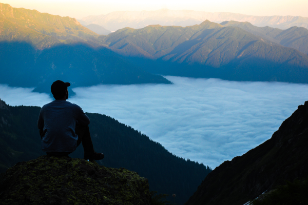 A man in a baseball cap contemplating clouds in a valley below him