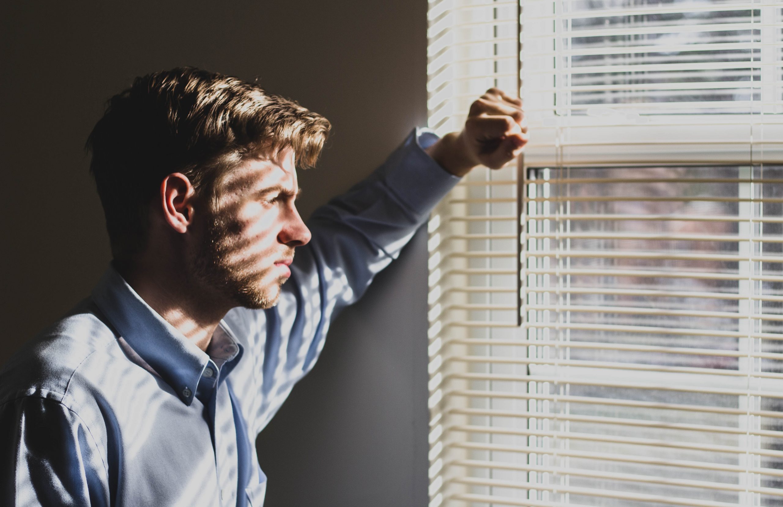 A man looking wistfully out of a window with blinds casting shadows on his face